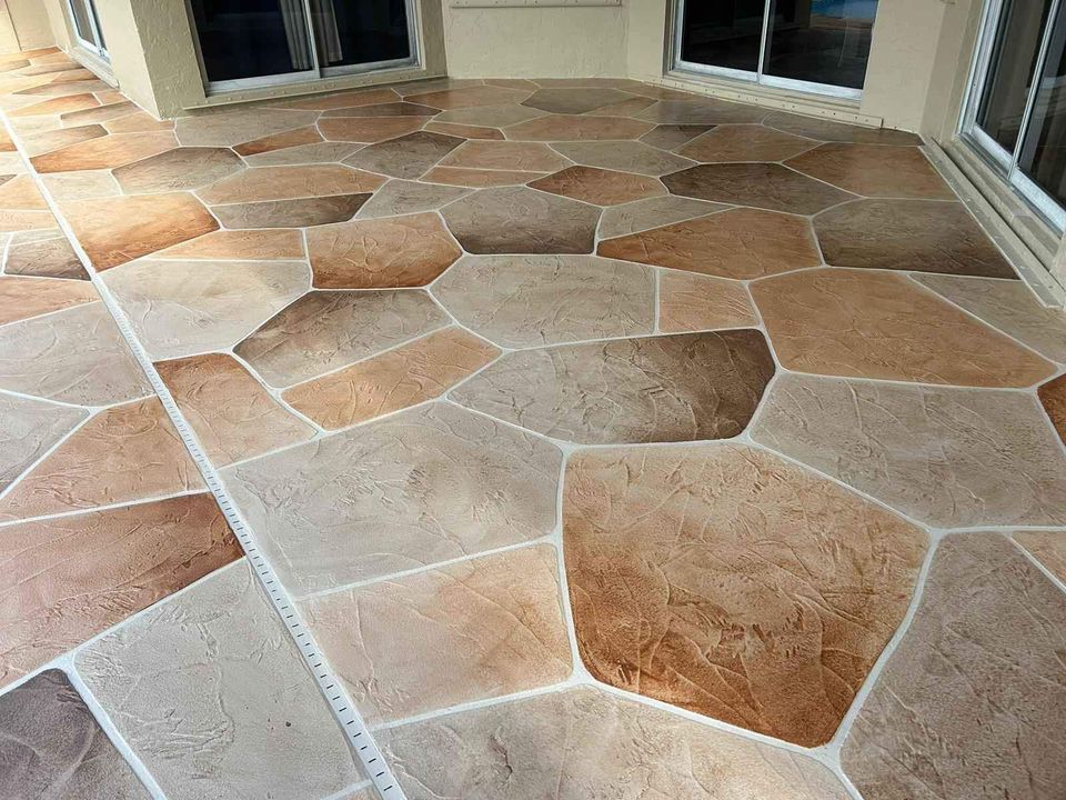 A patio floor featuring large, irregularly shaped tiles in shades of beige, tan, and brown, creating a natural stone appearance. The surface is surrounded by glass sliding doors.