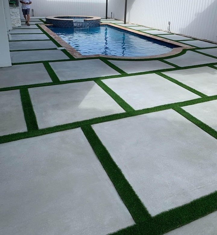 Modern backyard with a rectangular pool and hot tub. The patio has large square concrete slabs with artificial grass in between, creating a geometric pattern. A white fence encloses the area. One person stands near the pool.