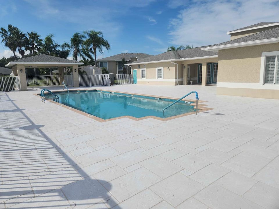 A rectangular outdoor swimming pool with metal handrails, surrounded by a spacious white tiled deck, next to a beige building under a blue sky with scattered clouds and palm trees in the background.