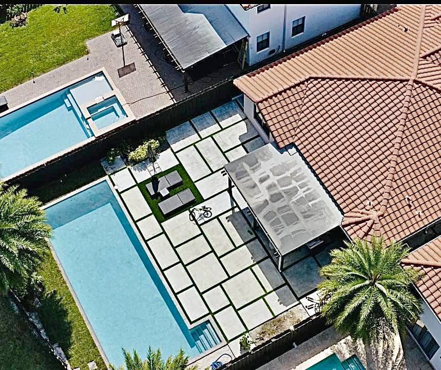 Aerial view of a modern house with a red-tiled roof, a large swimming pool, a shaded patio, palm trees, and a geometric paved yard. Two smaller pools are also visible in the surrounding area.